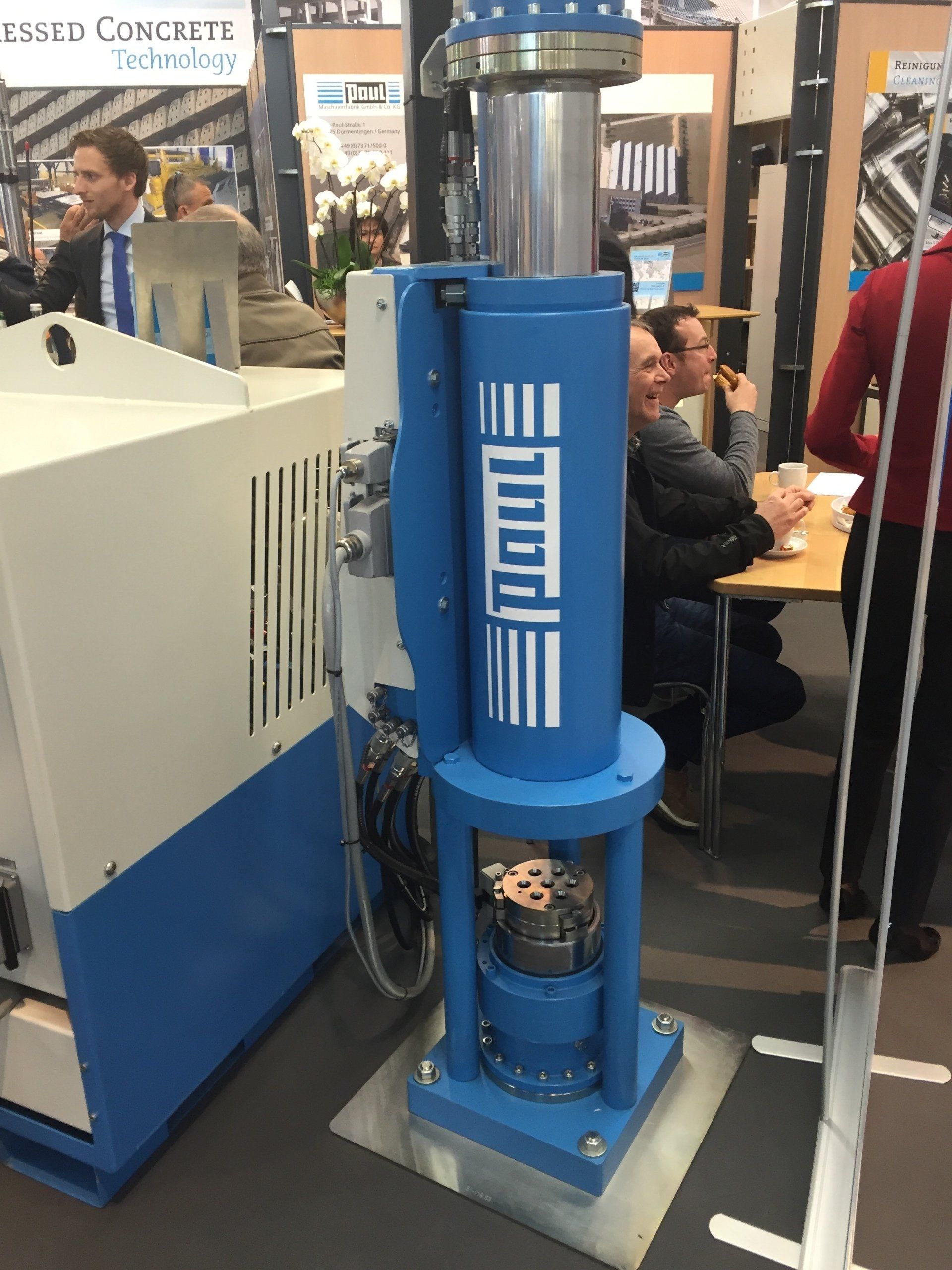 Blue industrial machine on display at a trade show, with people and booths in the background.