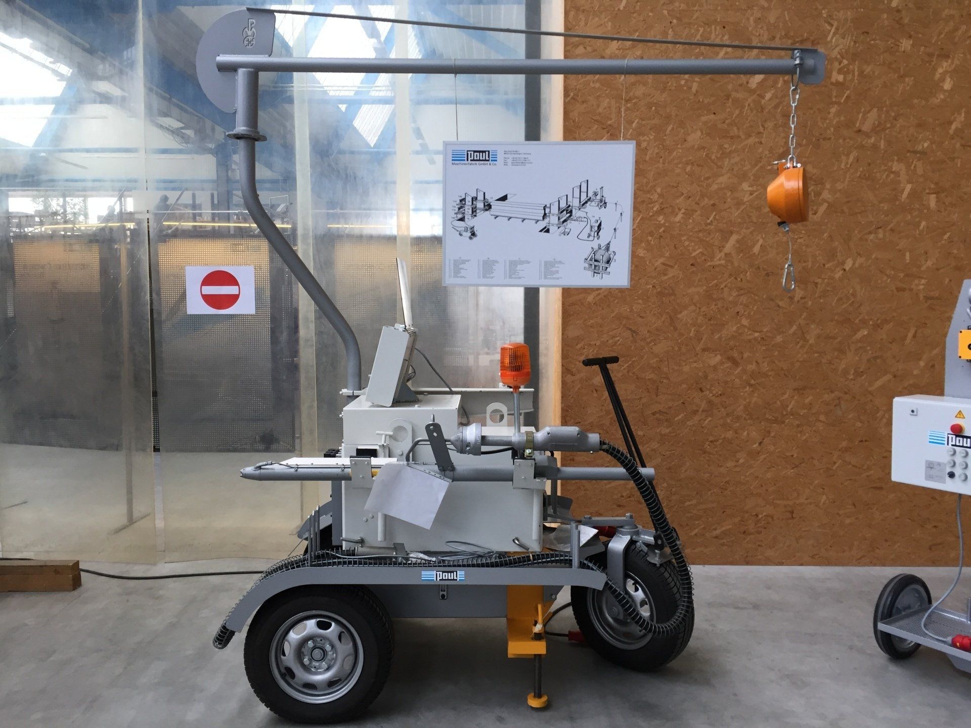Small mobile machine with an orange scoop in an indoor workshop, mounted on a trailer with a sign above it.