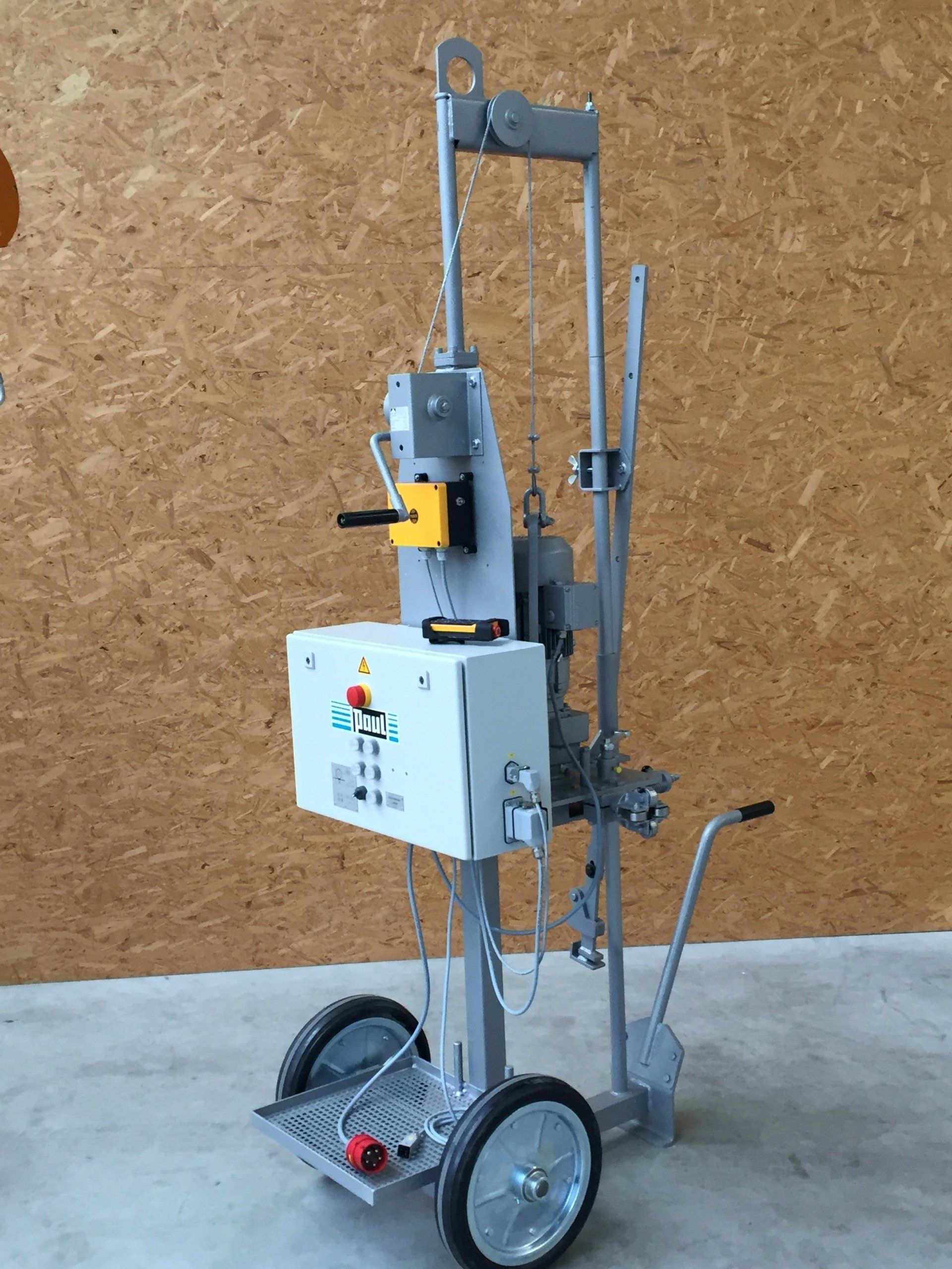 Industrial lifting machine with gray frame, control box, and wheels against a corkboard wall.