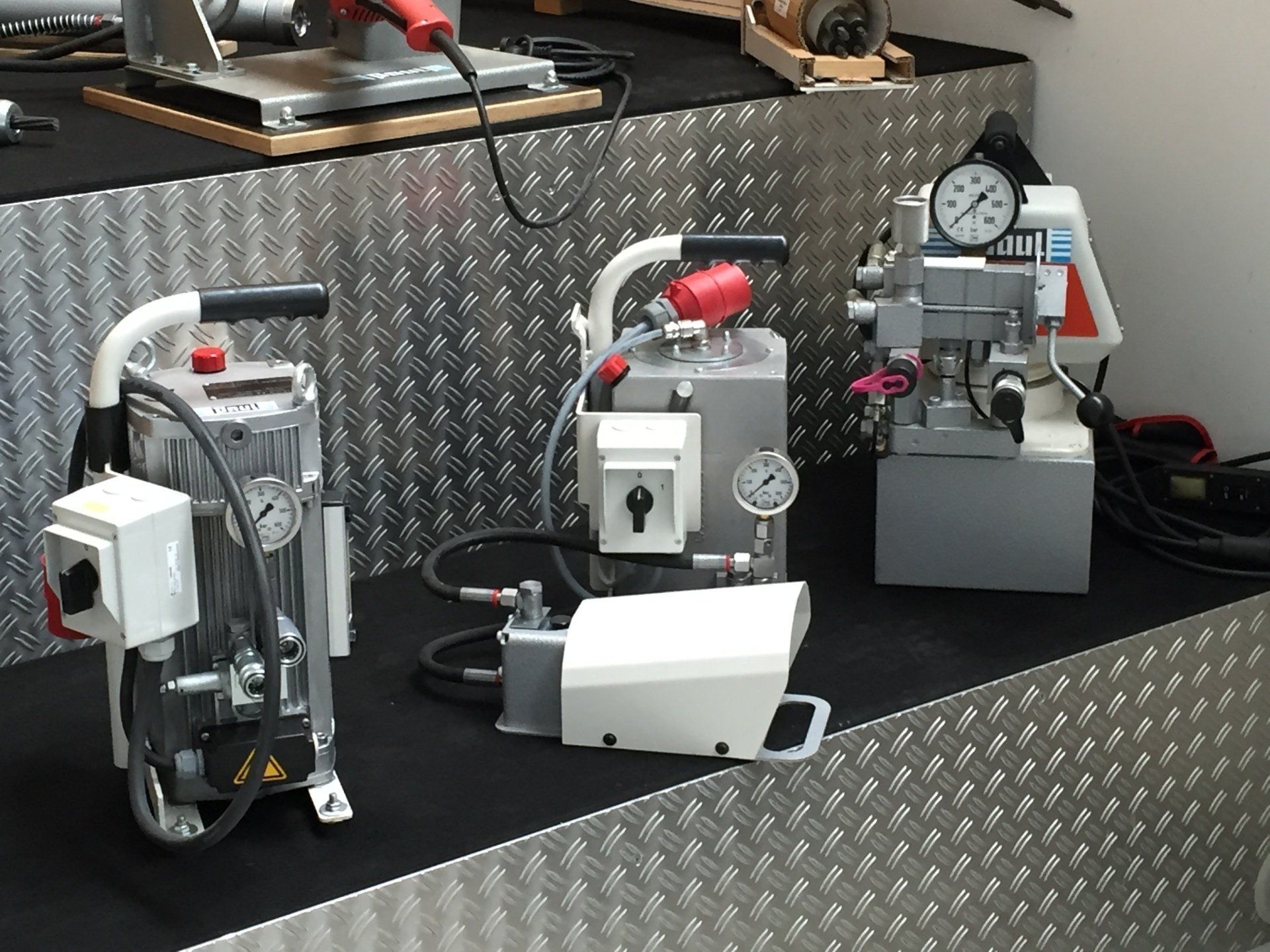 Two industrial machines with red knobs on a black workbench in a workshop, against a diamond-plate metal wall.