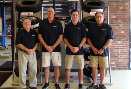Four men in black polo shirts and khaki shorts standing in a tire shop, smiling at the camera.