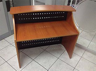 Mostrador de recepción de madera con panel posterior perforado, colocado sobre un suelo de baldosas.