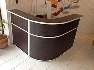 Mostrador de recepción curvado de color marrón oscuro con detalles en blanco, frente a una pared con ilustraciones de salón.