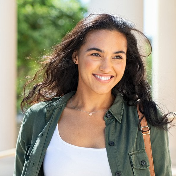 Woman with curly dark hair, smiling, wearing a green jacket over a white top, outside.