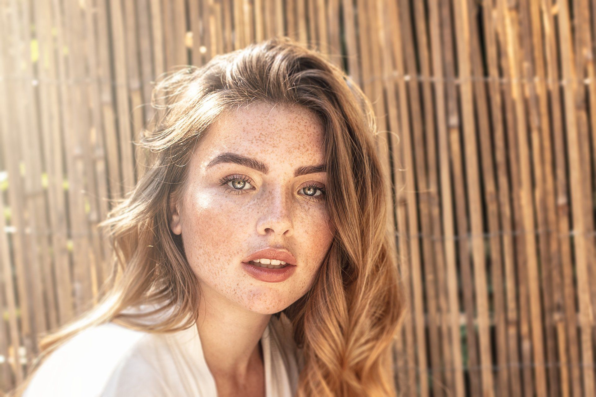 Woman with freckles and wavy blonde hair, looks at camera in front of a bamboo background.