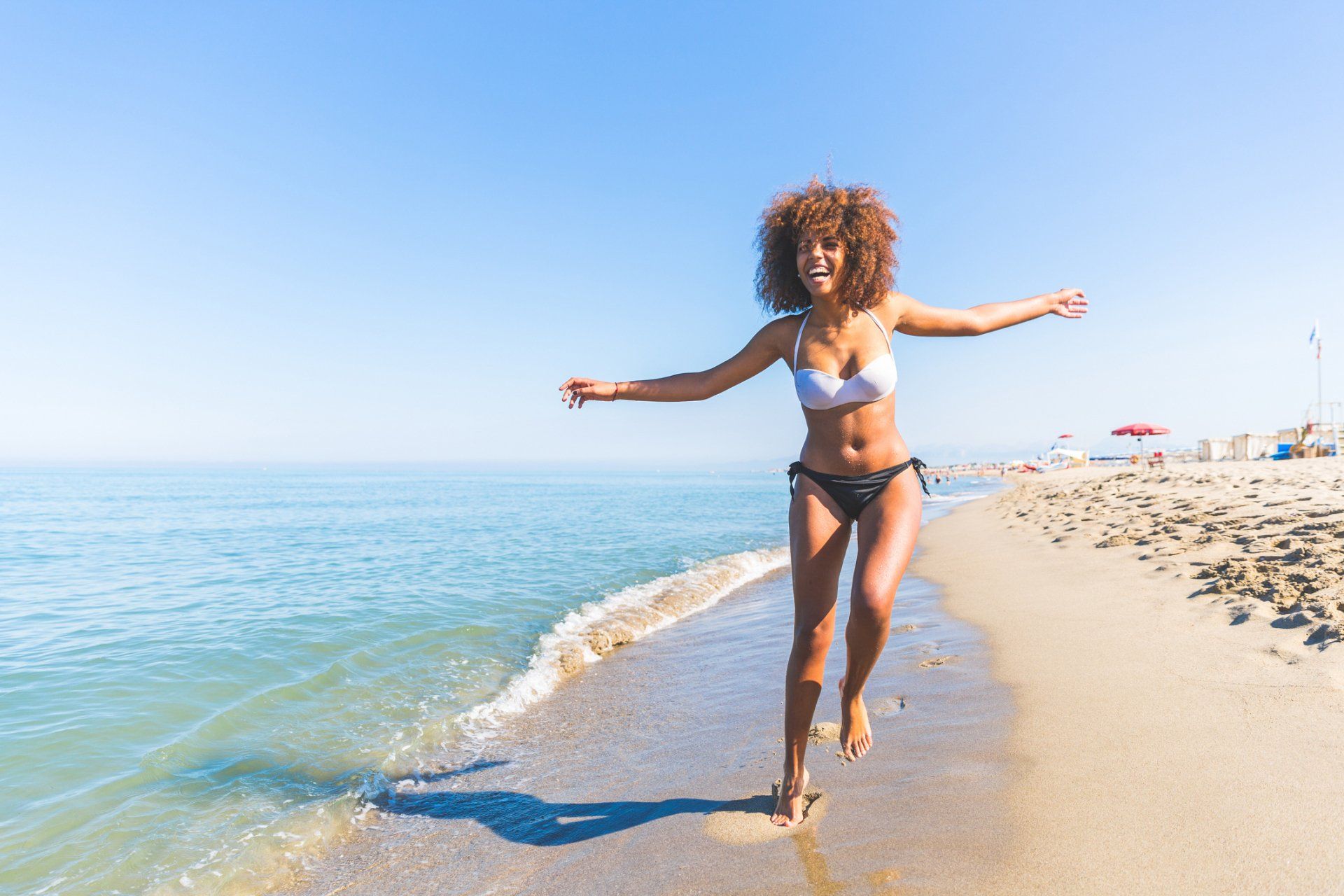 Woman in bikini runs happily along a sandy beach near the water, arms outstretched, under a blue sky.