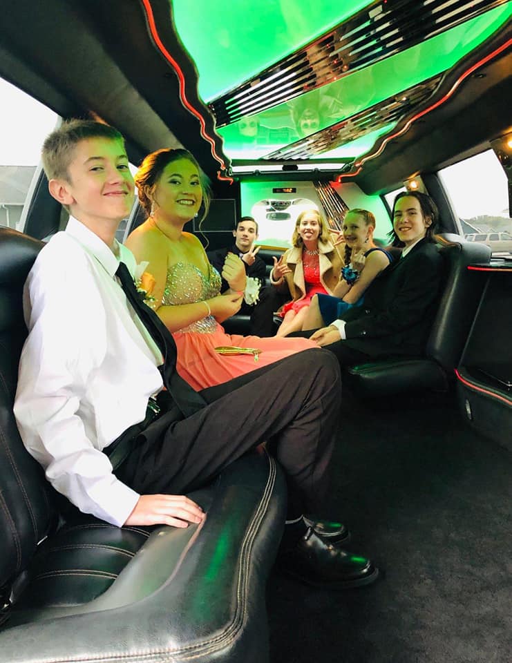 A group of six young adults, dressed in formal attire, sit inside a limousine with colorful lighting. They appear to be enjoying themselves, with some smiling and others gesturing peace signs—truly embracing the joy of celebrating your birthday being driven in a limo.