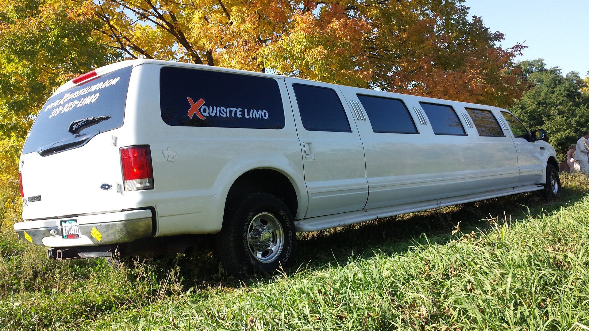 A white stretch limousine with dark tinted windows is parked on grass near trees, with a logo on the side reading 