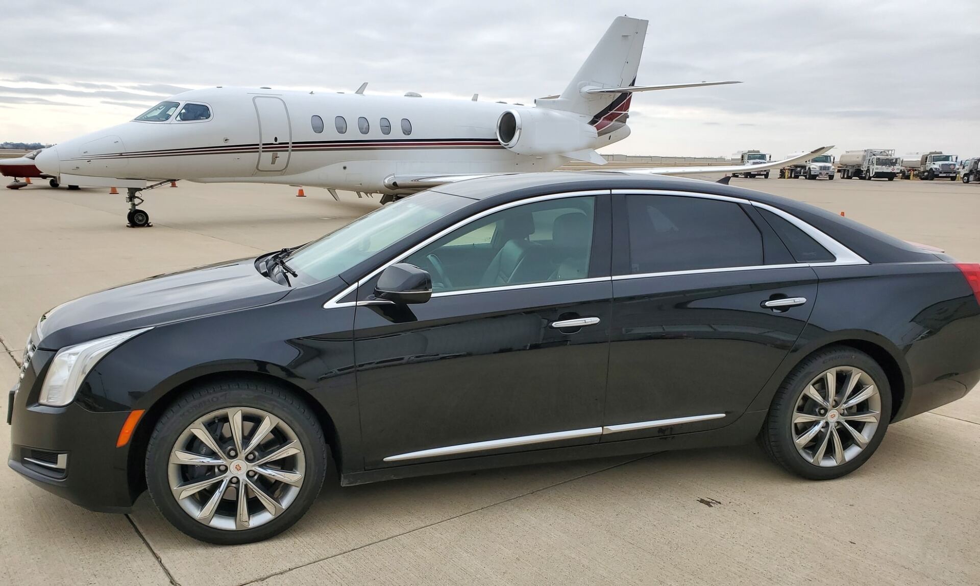 A black luxury sedan is parked on an airport tarmac with a white private jet in the background under a cloudy sky at Signature CID Cedar Rapids, IA