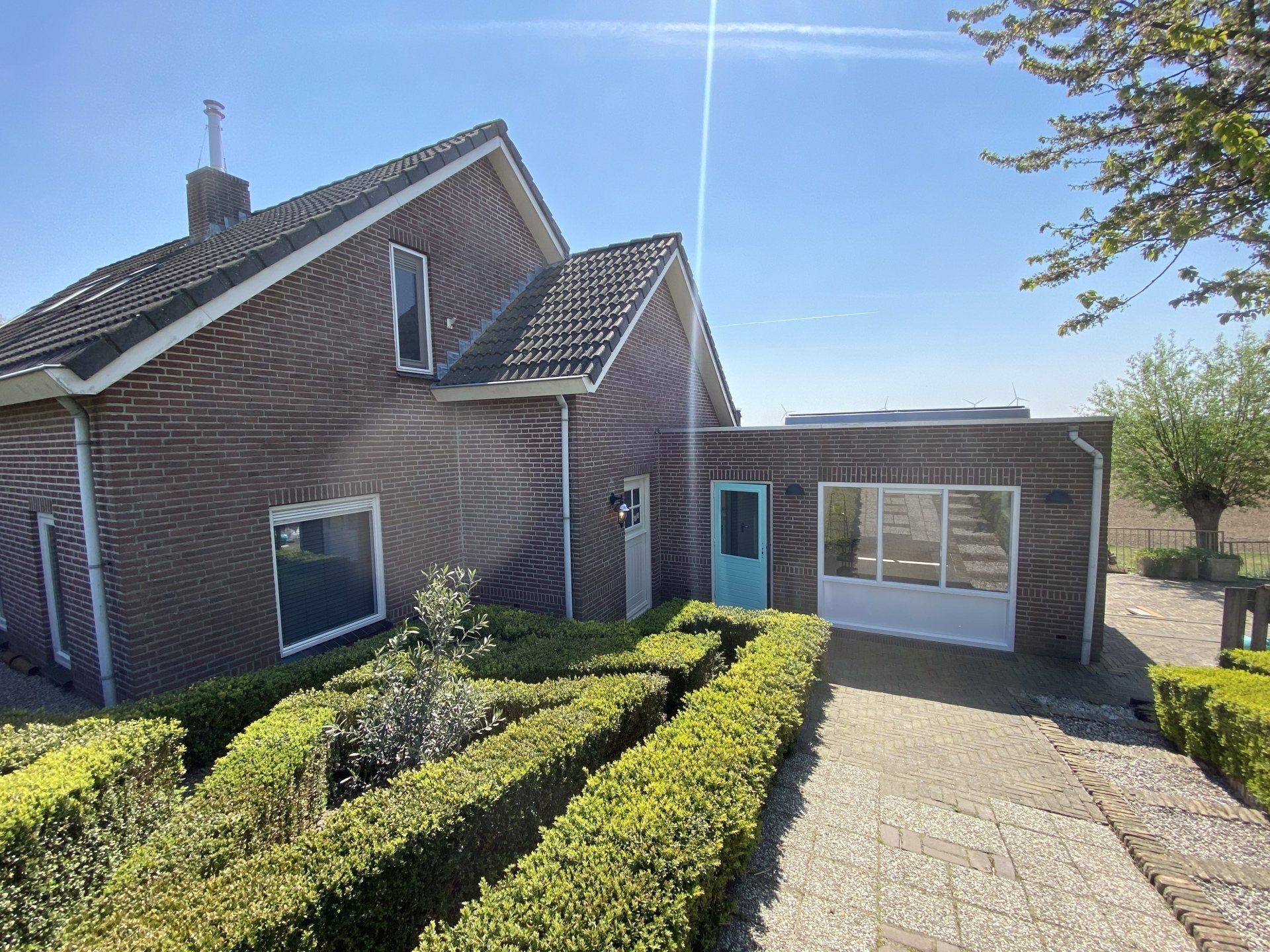Bakstenen huis met turquoise deur en aangebouwde constructie; grindpad en hegtuin ervoor.
