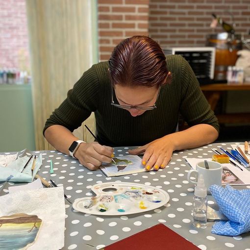 Louise schildert aan een tafel; palet en penselen zichtbaar. Groen shirt, bril. Tafelkleed met stippen.