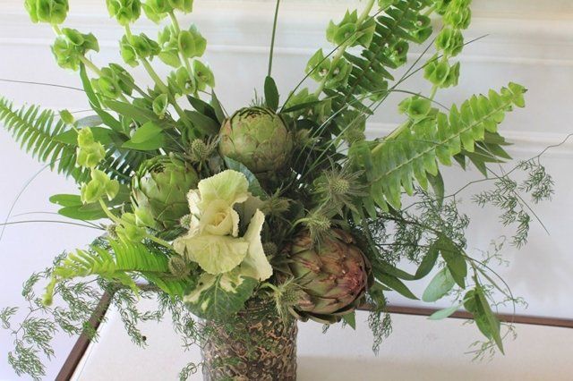 Green floral arrangement with artichokes, ferns, and other foliage in a vase.