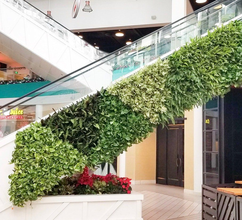 Escalator with a green wall of plants in a mall. A planter with red flowers sits at the base.
