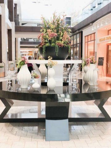 Large floral arrangement on black table in a mall, surrounded by white vases.