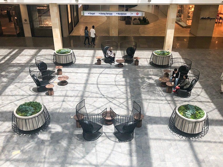 Overhead view of a mall seating area. Black chairs and tables, surrounded by large planters, sit on a gray floor.