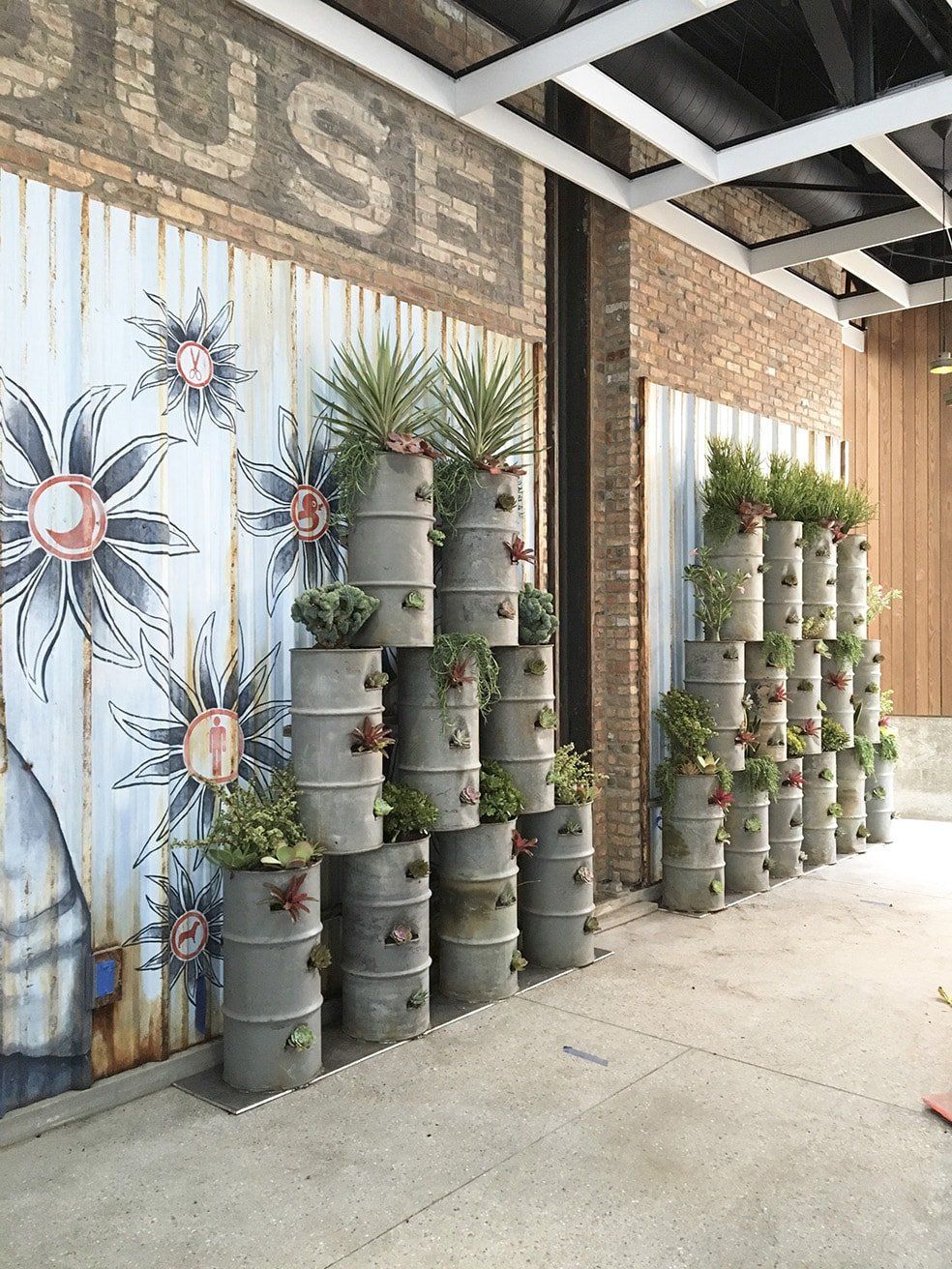 Vertical garden of stacked, gray barrels with plants against a wall with mural and exposed brick.