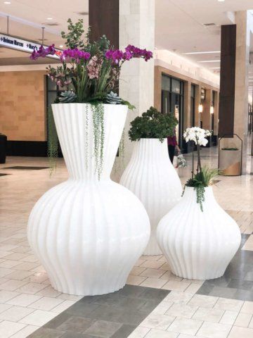 Three large, white, ribbed vases with colorful flowers and greenery in an indoor mall setting.