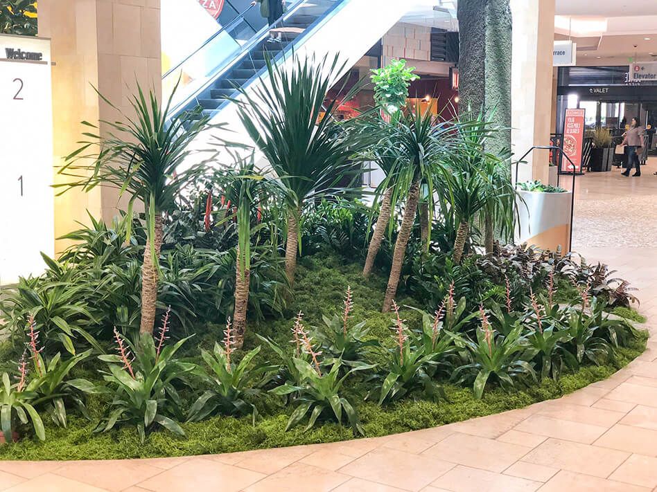 Indoor garden with palm-like plants at a mall. Escalator and signs in the background.