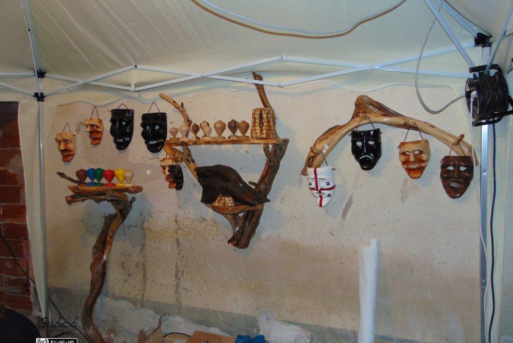 Una mostra di maschere fatte a mano e oggetti artigianali in legno appesi a strutture di legno galleggiante sotto un tendone bianco.