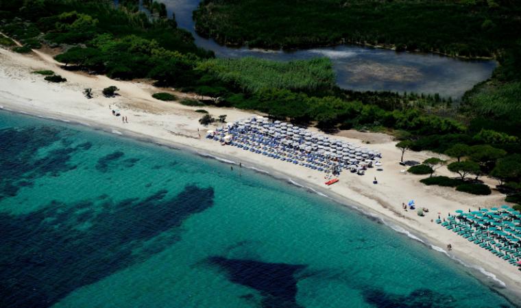 Veduta aerea di una spiaggia sabbiosa con ombrelloni blu, acqua turchese e un lussureggiante paesaggio costiero verdeggiante.