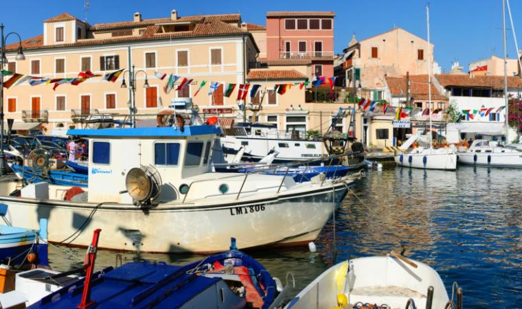 Barche ormeggiate in un soleggiato porto mediterraneo con edifici colorati e bandiere sullo sfondo.