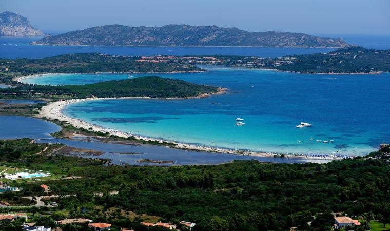 Una veduta dall'alto di una baia turchese con una spiaggia di sabbia bianca, orlata da una rigogliosa vegetazione verde e da una grande isola.