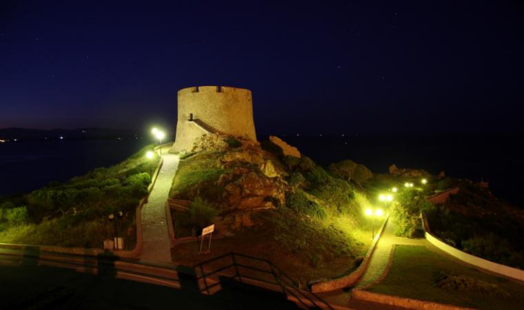 Una storica torre in pietra illuminata di notte, situata in cima a una collina erbosa con sentieri illuminati che conducono ad essa.