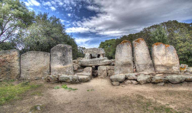 Una tomba dei giganti in Sardegna, caratterizzata da un muro curvo di grandi pietre verticali erose dal tempo, sotto un cielo azzurro nuvoloso.
