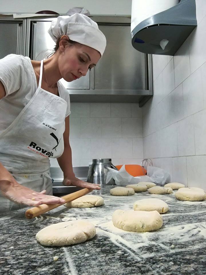 In una cucina, una persona con cappello e grembiule da chef usa un mattarello per stendere porzioni di impasto su un piano di lavoro in marmo.