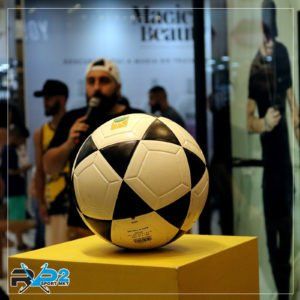 Bola de futebol em um suporte amarelo com um homem segurando um microfone na frente de uma multidão.