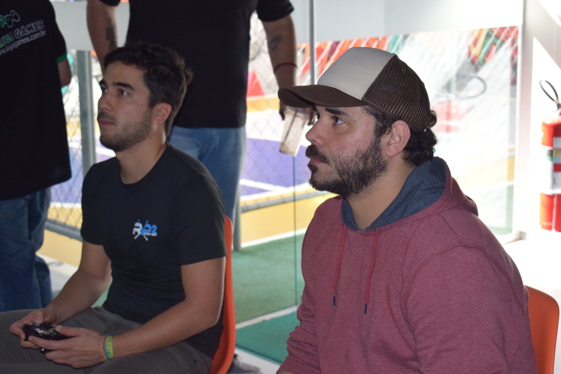 Dois homens jogando videogame; um usa boné e moletom vermelho, o outro, camiseta preta e segura um controle. Eles estão em uma sala bem iluminada.