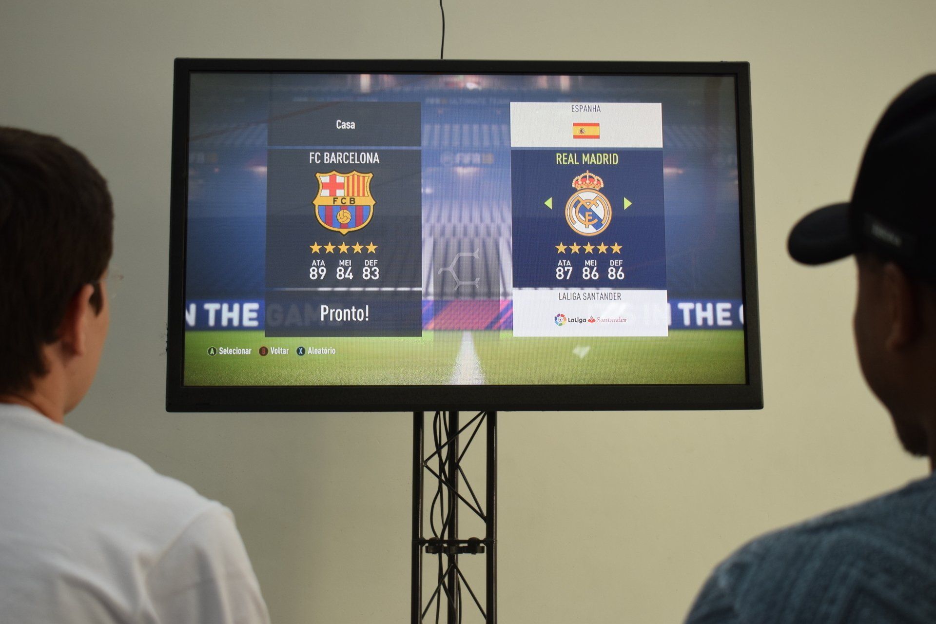 Duas pessoas jogando futebol, selecionando os times FC Barcelona e Real Madrid em um monitor grande.