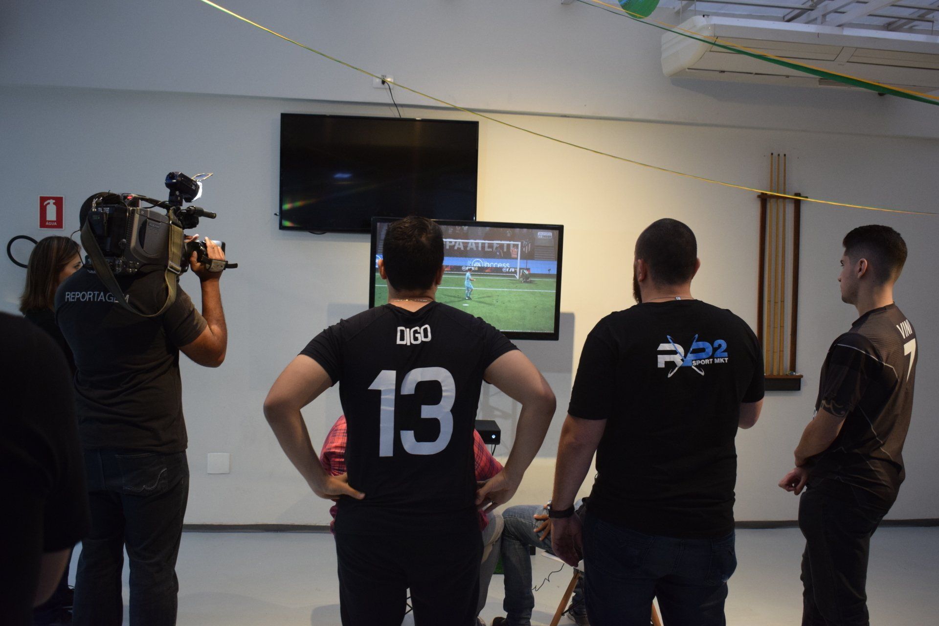 Pessoas assistem a uma partida de futebol na TV. Um cinegrafista filma a cena. O cenário parece ser um escritório ou estúdio.