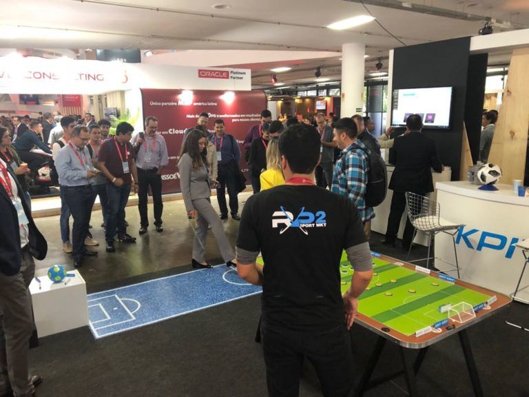 Pessoas assistem a uma partida de futebol de robôs em um evento empresarial. Um homem de camisa preta está em pé em frente à mesa de jogo, observando.
