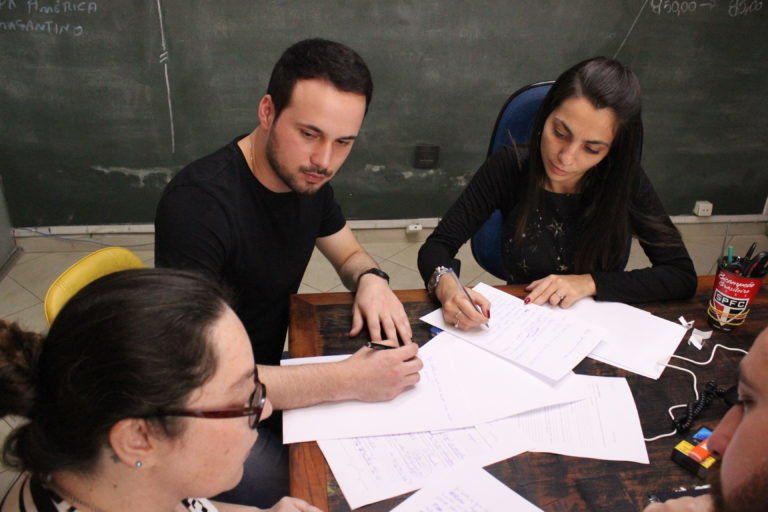 Quatro pessoas sentadas ao redor de uma mesa, trabalhando em papéis. Um homem e uma mulher estão concentrados, os outros dois parcialmente visíveis. Interior escuro, possivelmente uma sala de aula.