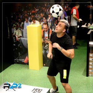Um homem de roupa preta equilibra uma bola de futebol na cabeça. Ele está em pé sobre uma superfície semelhante a grama, em frente a uma parede de pessoas.