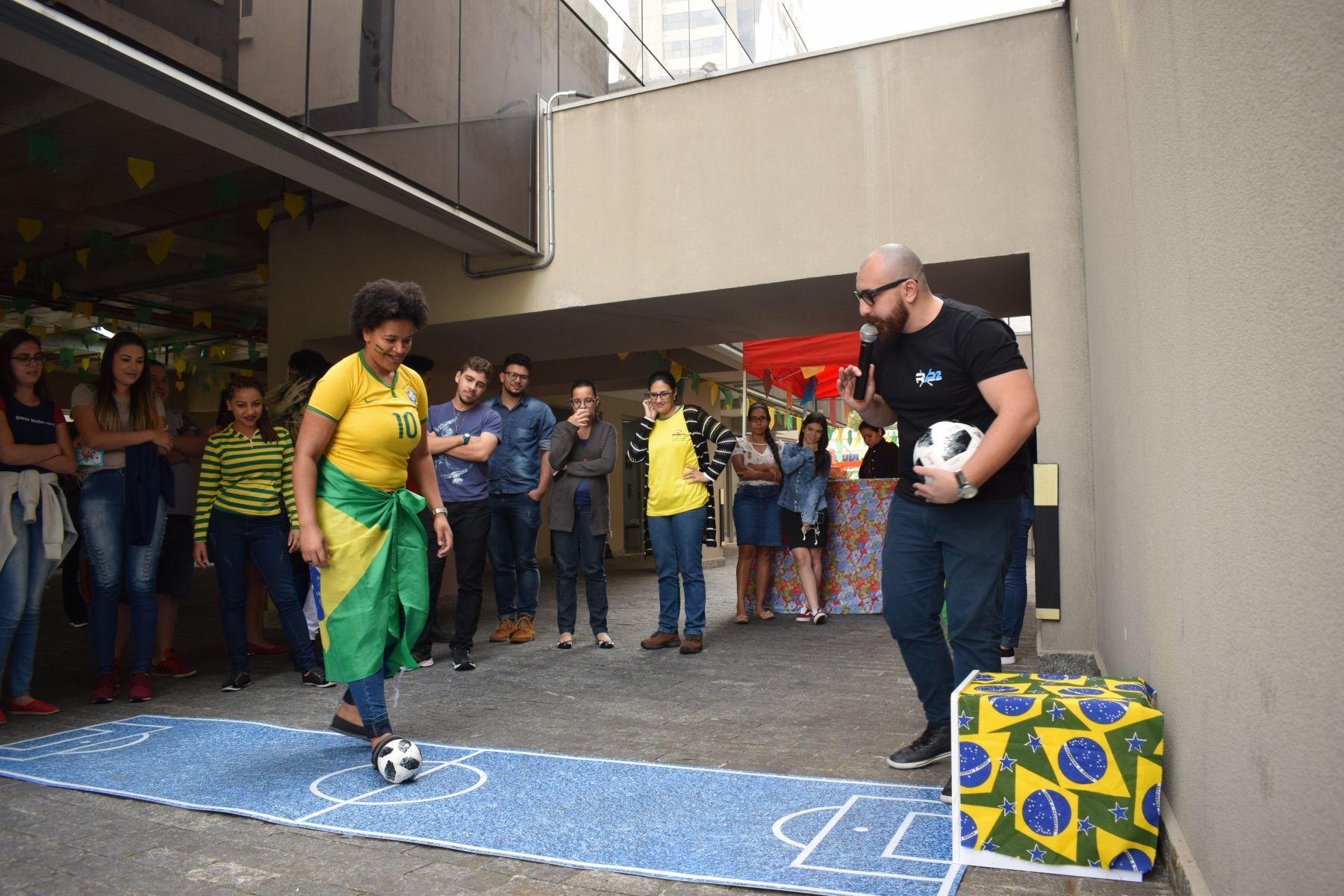 Mulher de amarelo e verde chuta uma bola de futebol em um campo pintado de azul, observada por uma multidão; um homem segura uma bola por perto.