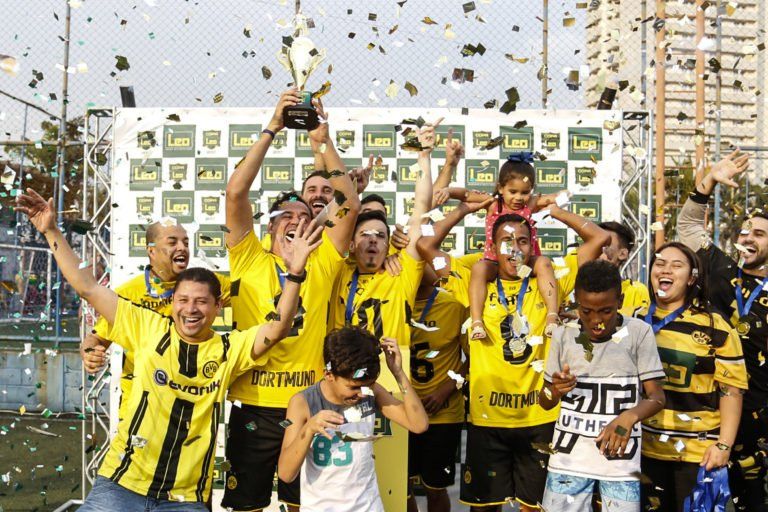 Time de futebol com uniforme amarelo e preto comemorando a vitória no campeonato, segurando um troféu com confete caindo.