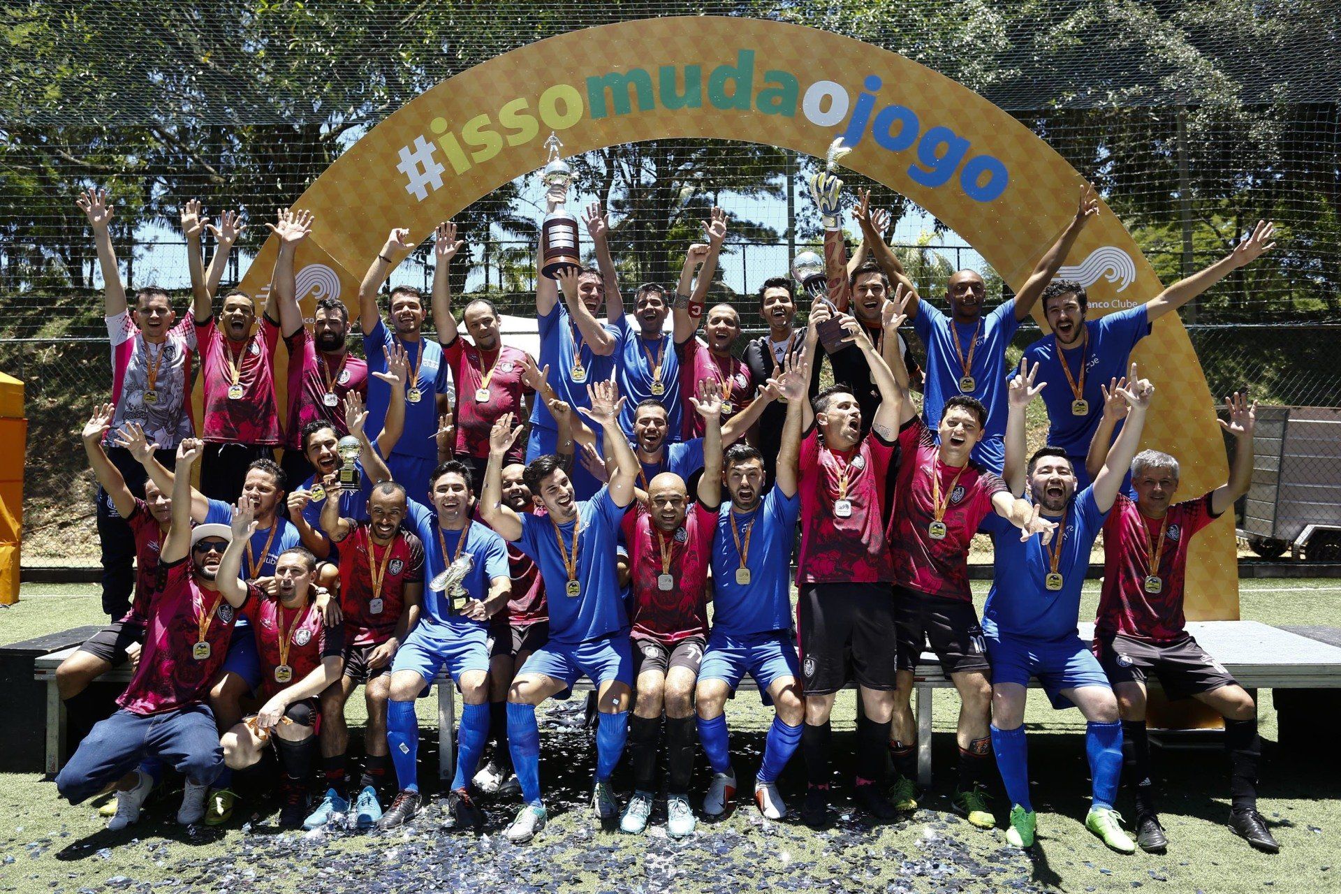 Grupo de jogadores de futebol, vestindo camisas azuis e vermelhas, comemorando uma vitória ao ar livre. Eles erguem os braços e seguram um troféu sob um arco.