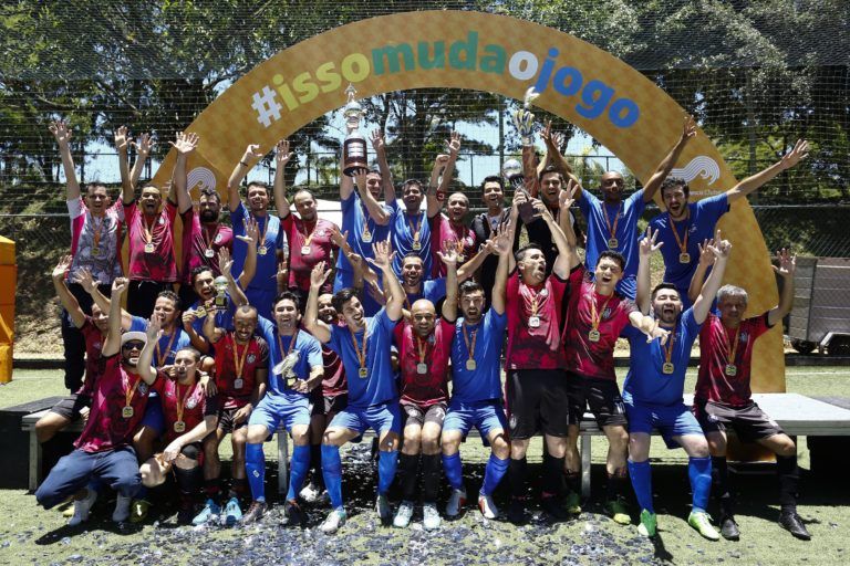 Um time de futebol, vestindo camisas azuis e vermelhas, comemora uma vitória ao ar livre, erguendo um troféu e os braços para o alto. Eles estão em frente a um arco com a inscrição 