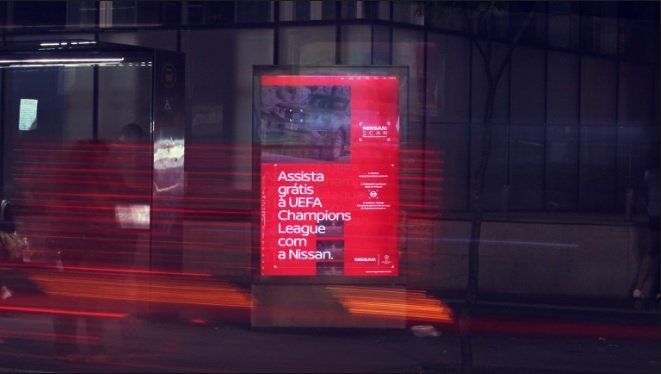 Outdoor noturno anunciando a Liga dos Campeões da UEFA em vermelho e branco com desfoque de movimento causado por luzes que passam.