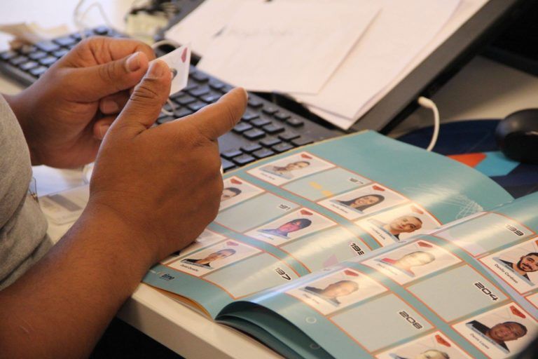 Pessoa segurando um pequeno cartão, olhando para um livro aberto com cartões de fotos, presumivelmente uma coleção ou álbum, sobre uma mesa com um teclado.