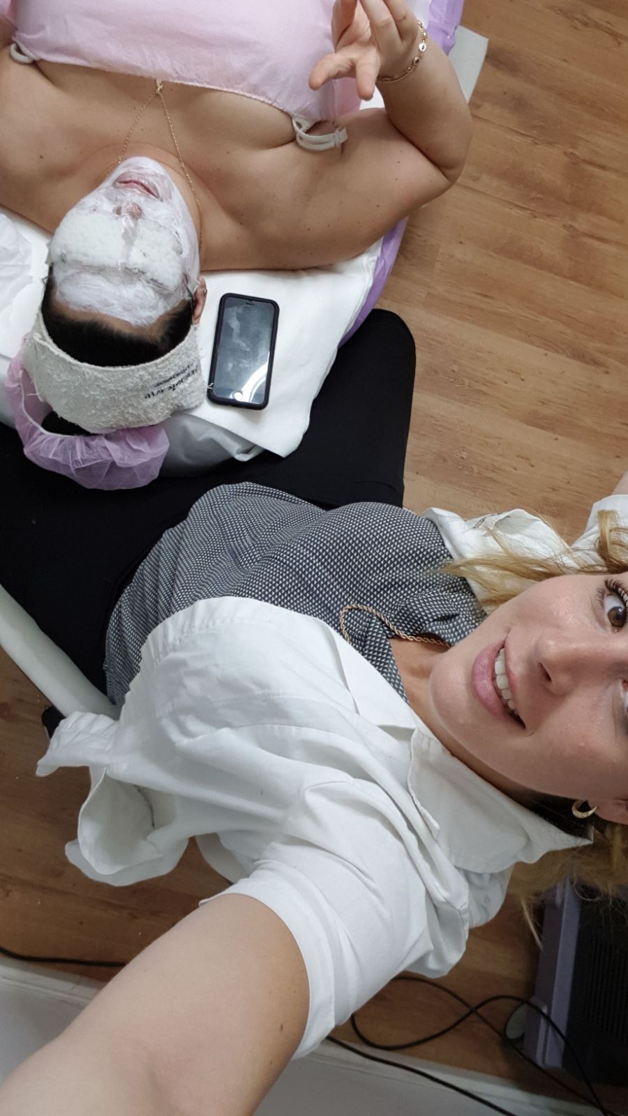 Two people lying on a wooden floor, one taking a selfie near a phone and bag.