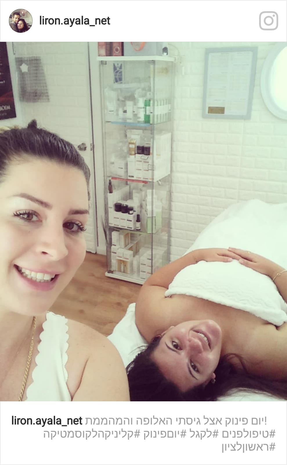 Two women in a bright salon, one taking a selfie while another lies back with a white towel on her face.