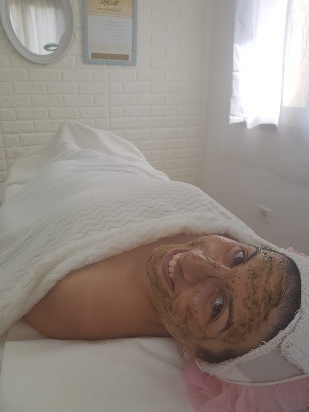 Person lying in bed under a white blanket, smiling in a bright bedroom near a window.