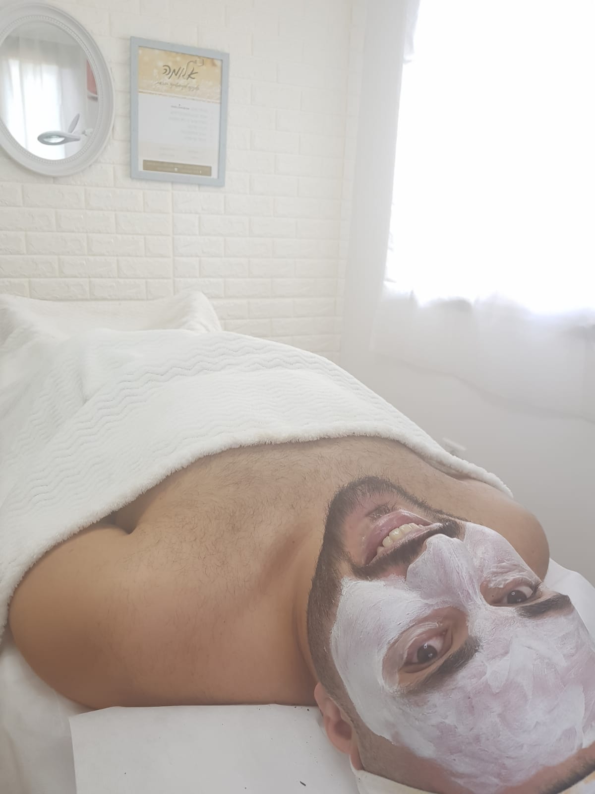 Person lying on a massage table with a white facial mask in a bright spa room