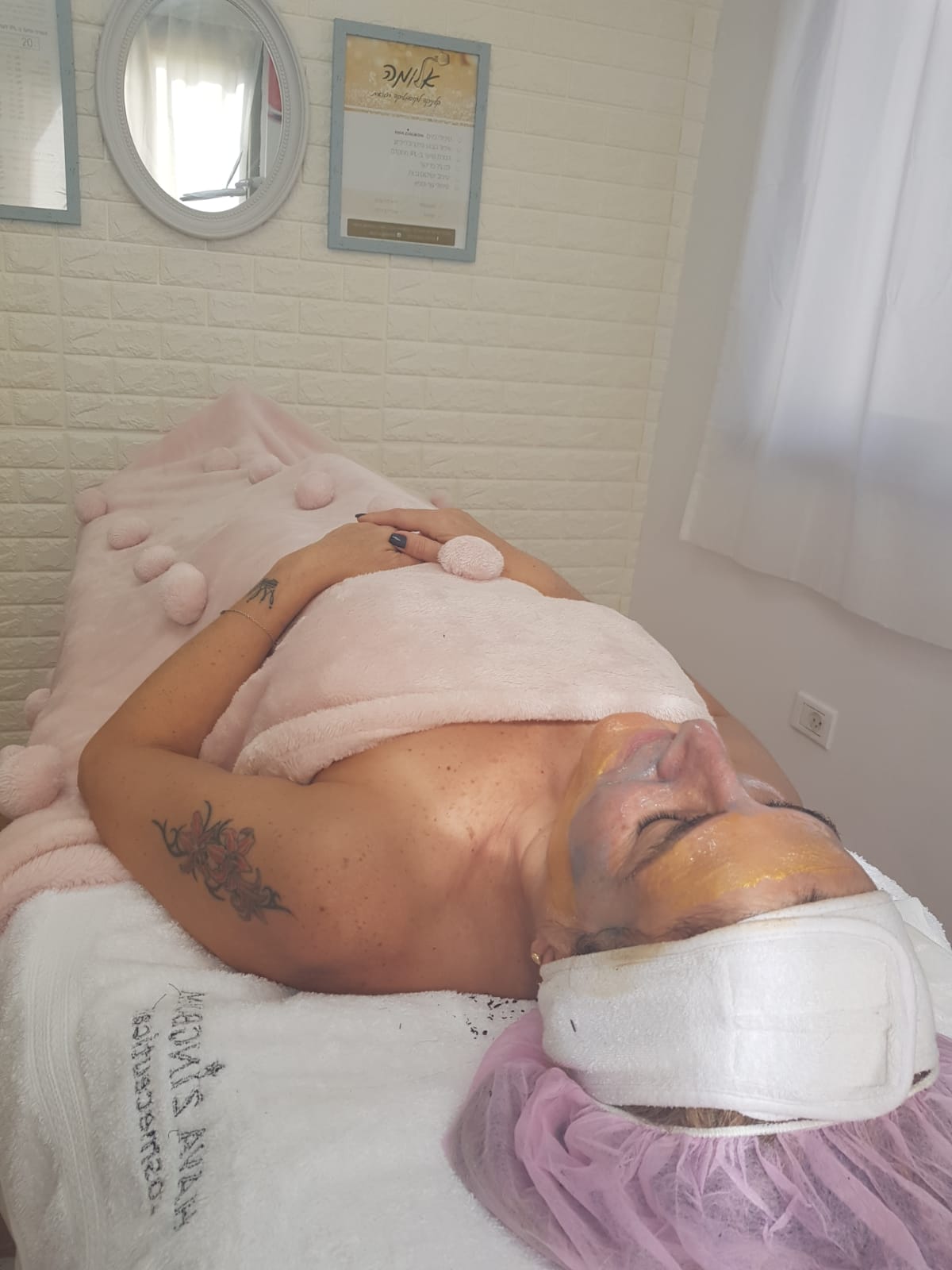Person lying on a spa treatment table with a white towel and pink head wrap in a soft, serene room