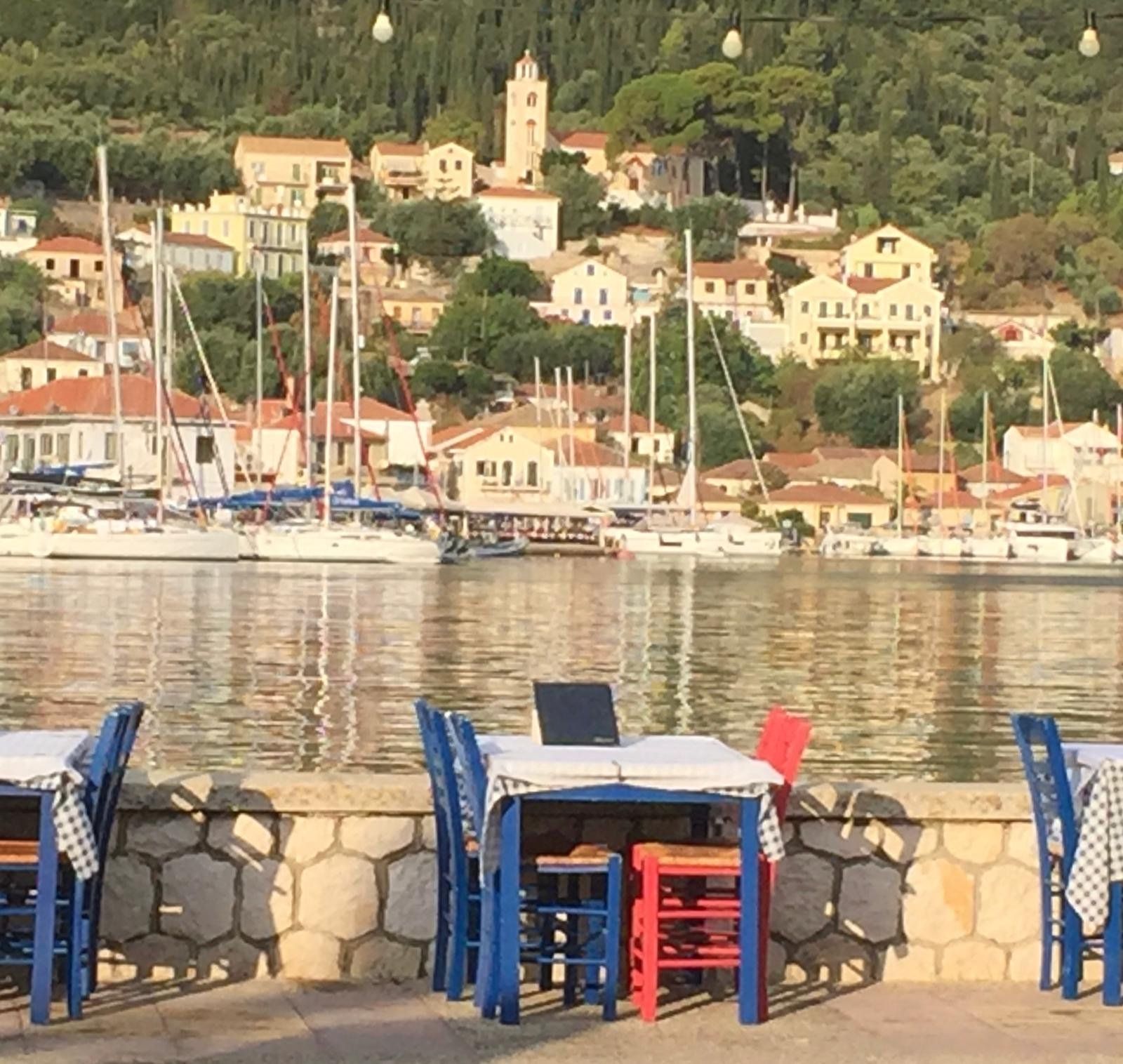 Waterfront café tables and blue chairs facing a calm harbor with moored sailboats and hillside buildings.