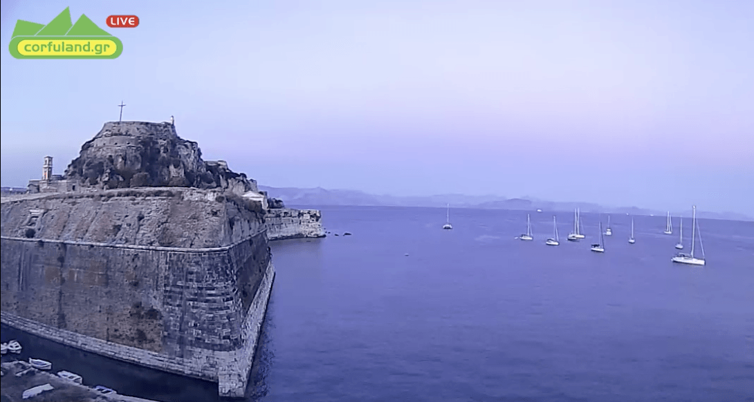 Webcams Corfu Stone fortress on a cliff overlooking a calm blue sea with several sailboats at sunset.