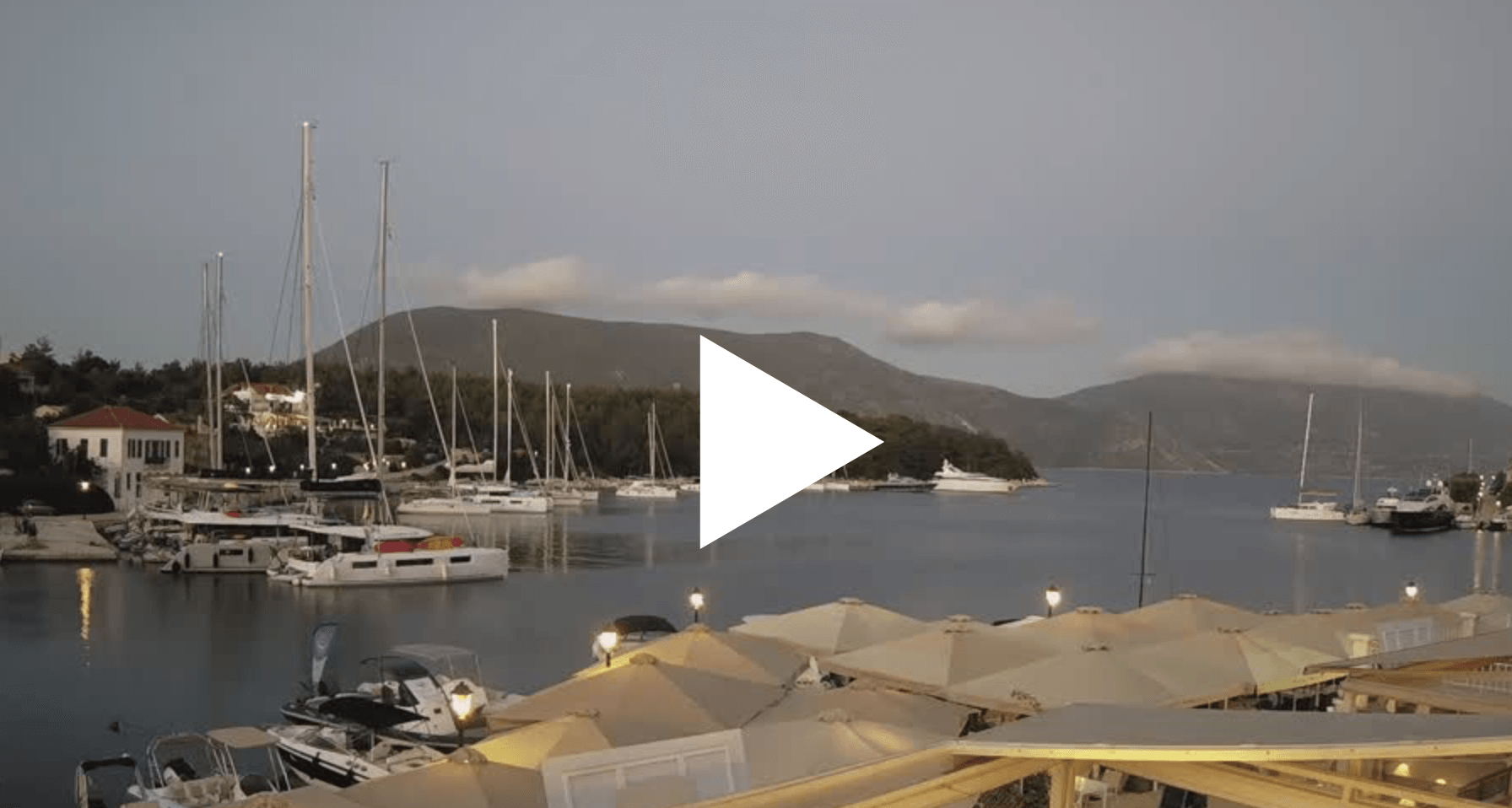 Webcam Fiskardo Marina with sailboats at sunset, mountains in the distance, and a play button overlay.